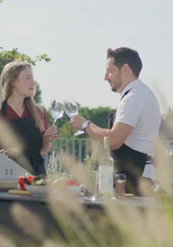 Ein Mann und eine Frau stehen draußen in einer Außenküche, kochen und trinken dabei Wein.