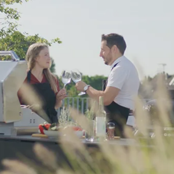Ein Mann und eine Frau stehen draußen in einer Außenküche, kochen und trinken dabei Wein.